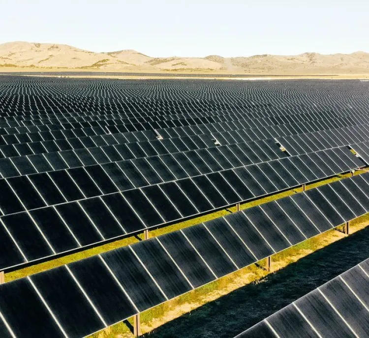 A vast solar farm with rows of solar panels stretching into the distance, set against a backdrop of rolling hills under a clear sky.