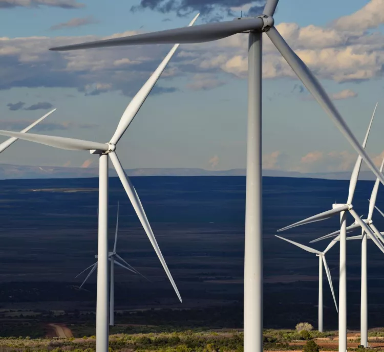 A series of wind turbines with large blades stand in an open landscape under a partly cloudy sky. The turbines are aligned in a row, stretching into the distance, with hills visible on the horizon.