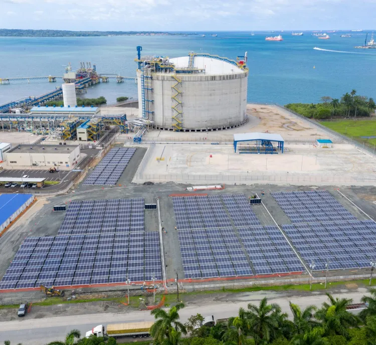 Aerial view of a coastal industrial area with a large circular storage tank, solar panels, and buildings near the ocean.