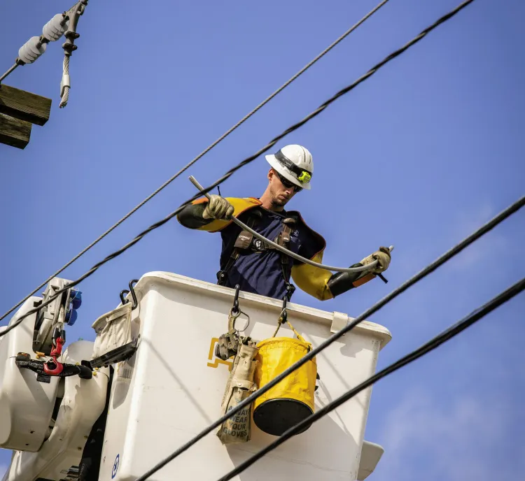 Utility worker in a bucket lift repairing power lines against a clear blue sky.
