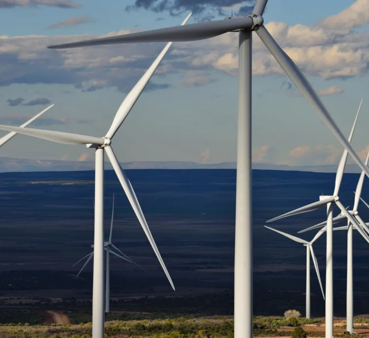 A row of wind turbines in a vast open landscape under a partly cloudy sky. The turbines are large with white blades, and the view extends to a distant horizon.