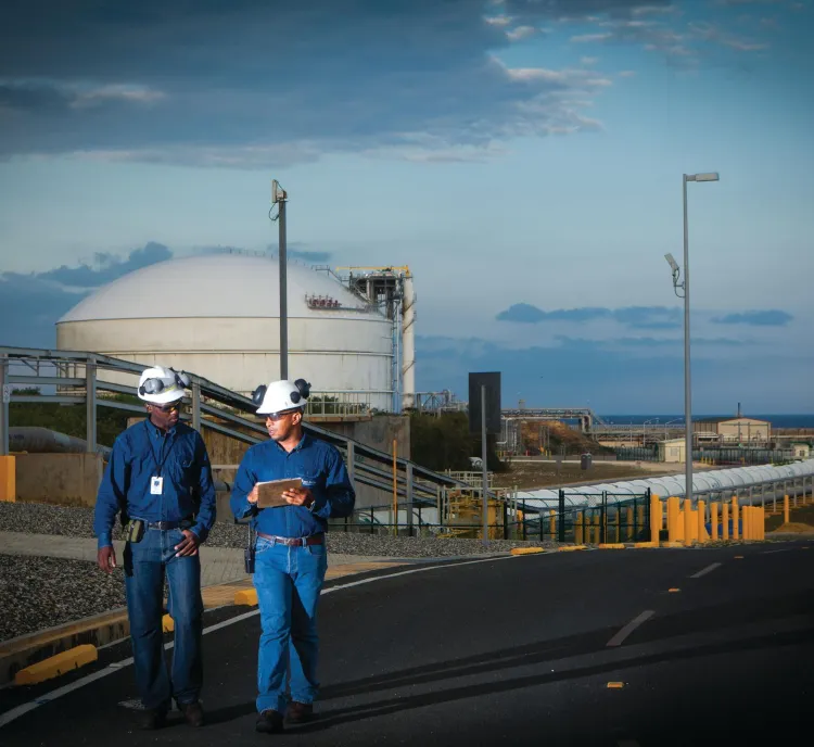 Two workers wearing blue uniforms and white hard hats walk along a road in an industrial area. They are holding a clipboard and appear to be in discussion. In the background, there is a large white storage tank and various industrial structures under a partly cloudy sky.