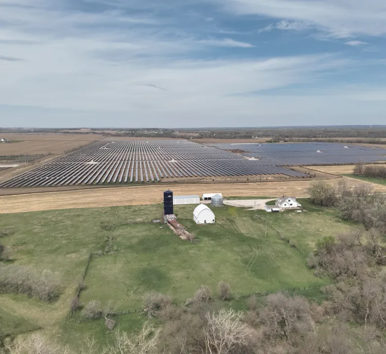 Aerial view of a large solar farm with rows of solar panels on a vast field. In the foreground, there is a small farm with a barn and silo surrounded by green fields and trees. The sky is partly cloudy.