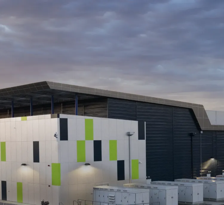 Modern industrial building with a geometric facade featuring green and gray accents, surrounded by rows of white utility cabinets, photographed at sunset under a cloudy sky.