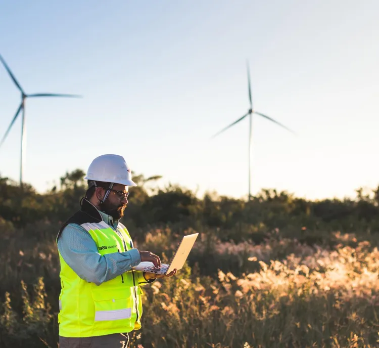 Engineer wearing a safety vest and hard hat uses a laptop in a field with wind turbines in the background during daylight.
