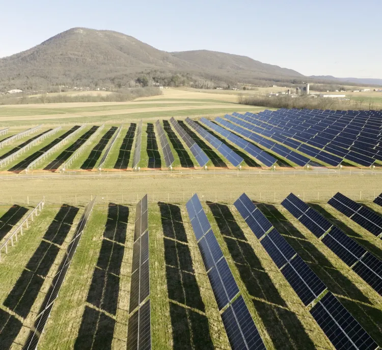 A large solar farm with rows of solar panels in a grassy field, set against a backdrop of rolling hills under a clear blue sky.