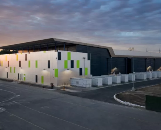 Modern industrial building with colorful panels and large roof at dusk, surrounded by containers and a paved area.