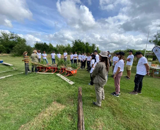 A group of people gathered outdoors for a workshop, standing on grass near wheelbarrows filled with plants. They are listening to a person speaking at the front. The sky is partly cloudy, and there are trees in the background.