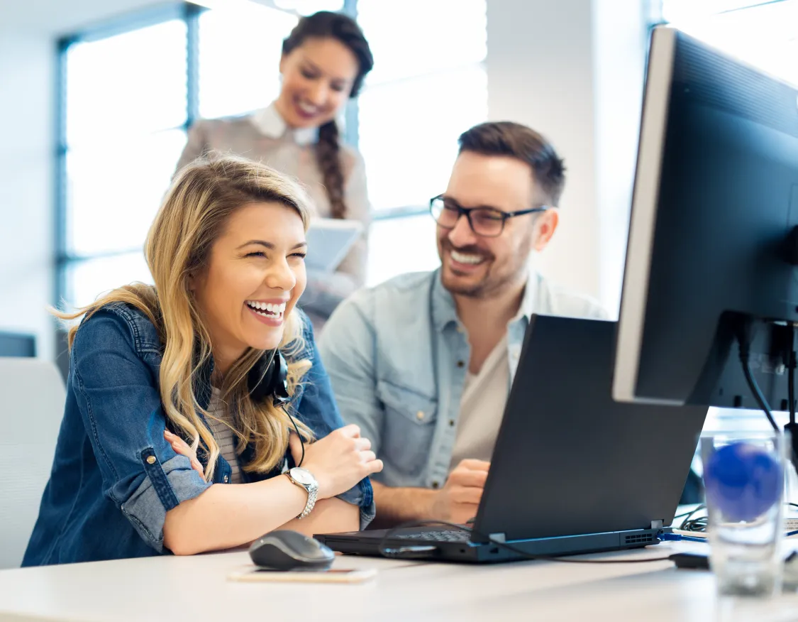 Three colleagues in an office laughing and looking at a computer screen. A woman in the foreground is smiling broadly, while a man next to her and another woman standing behind them are also smiling.