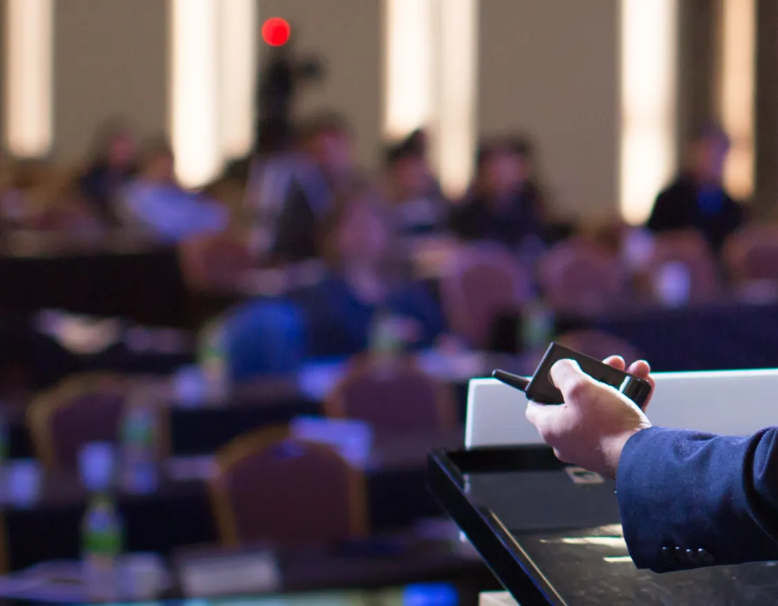 A person in a suit stands at a podium, holding a microphone, facing a blurred audience in a conference setting.