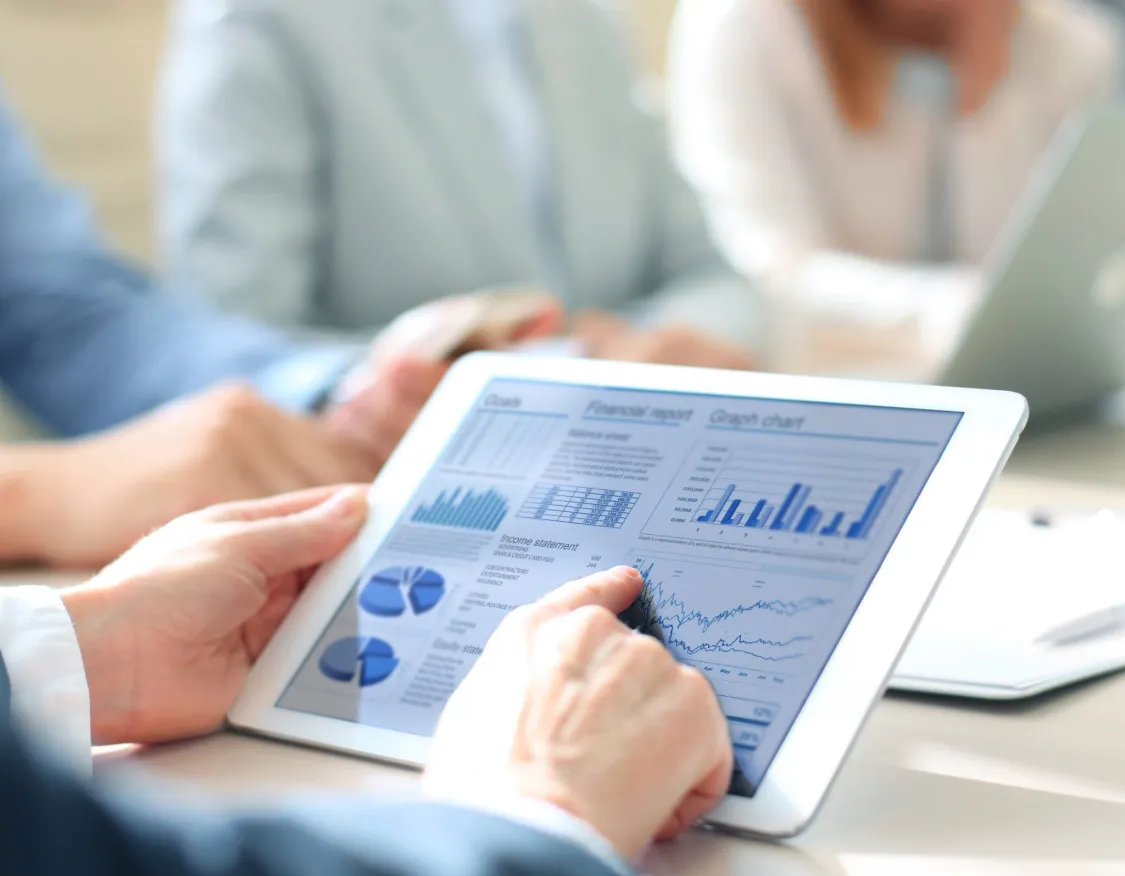 A person using a tablet to view financial reports with graphs and charts, surrounded by colleagues in a meeting.