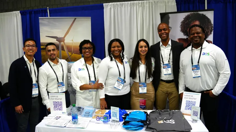 Seven people stand behind a table at a conference booth displaying AES branding. They are smiling and wearing badges. The table is covered with promotional items, brochures, and a sign encouraging visitors to leave their resume. Behind them are banners with images of a wind turbine and a person wearing headphones.