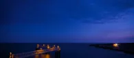 A long pier with lights extends into the dark blue sea under a twilight sky. The horizon is visible with a faint glow on the right.