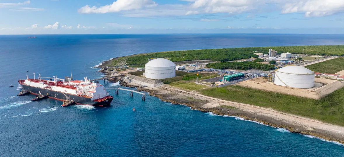 Aerial view of a coastal industrial area with two large white storage tanks near the shoreline. A large ship is docked at the pier, surrounded by clear blue water. The background shows green vegetation and a blue sky with scattered clouds.