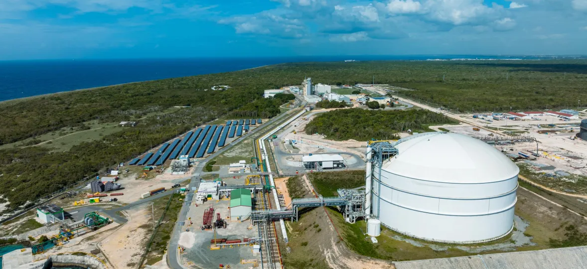 Aerial view of an industrial complex near the coast, featuring a large white storage tank, solar panels, and various buildings surrounded by greenery. The ocean is visible in the background under a partly cloudy blue sky.