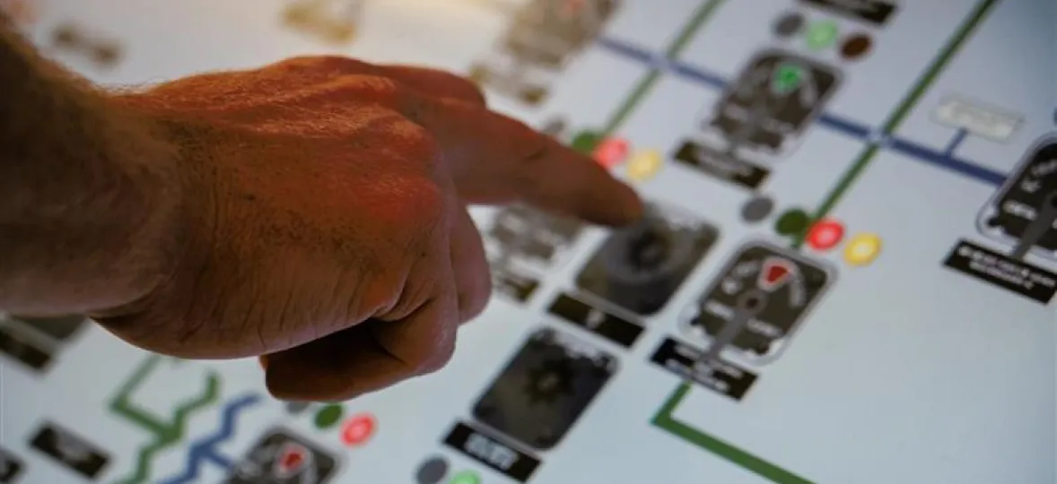 A person's hand is pointing at a complex control panel with various buttons and indicators. The panel features multiple colored lights and switches, suggesting a technical or industrial setting.