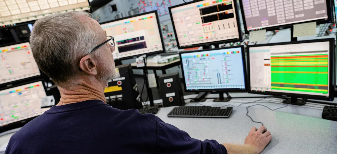 A person sits at a desk with multiple computer monitors displaying various data and graphs. The individual is using a mouse, focusing on the screens. The room appears to be a control center with a complex setup of technology.