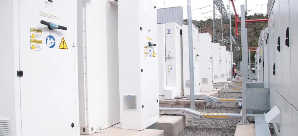 A row of white electrical cabinets with warning signs, including electrical hazard symbols, in an outdoor industrial setting. A person in a hard hat is visible in the background near power lines and metal structures.