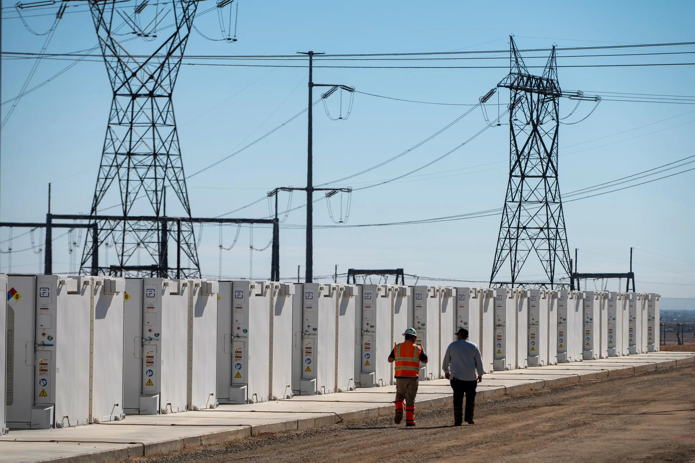 Two workers walk along a row of large white battery storage units at a power facility, with high voltage transmission towers and power lines in the background under a clear blue sky.