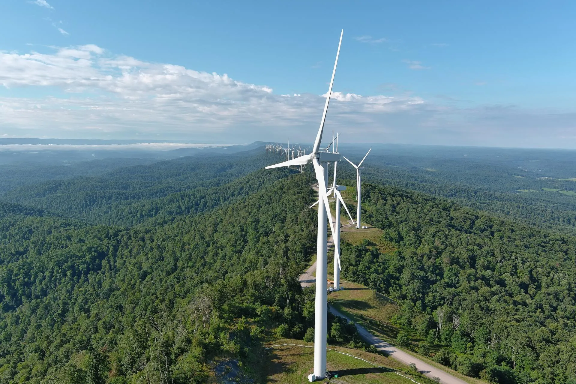 Aerial view of a row of wind turbines on a green forested ridge under a blue sky with scattered clouds.