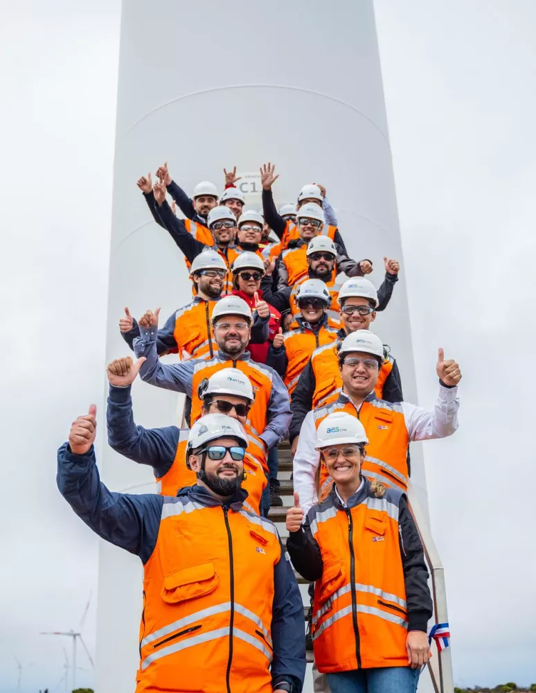 A group of people wearing orange safety vests and white helmets stand on a staircase in front of a wind turbine, giving thumbs up. The sky is overcast, and wind turbines are visible in the background.