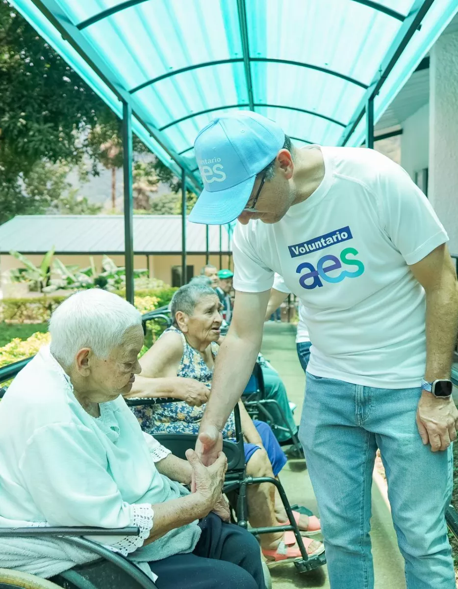 A volunteer in a white shirt and blue cap is gently holding the hand of an elderly woman in a wheelchair under a shaded walkway. Other elderly individuals are seated nearby, surrounded by greenery.