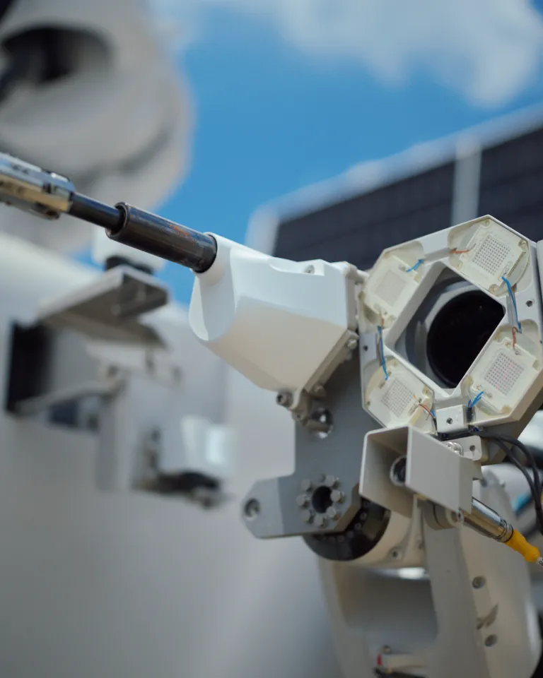 Close-up of a robotic arm with a metallic tool, featuring intricate wiring and components, against a blurred background with blue sky and clouds.