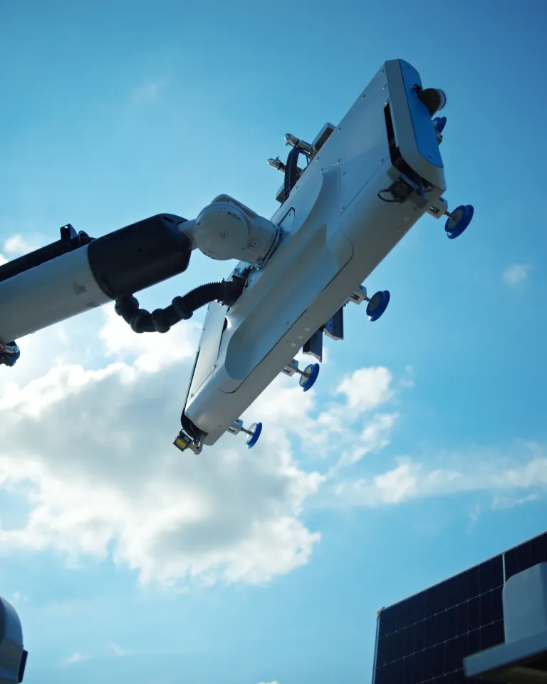 A robotic arm with suction cups reaches towards the sky, set against a backdrop of blue sky and clouds. A solar panel is partially visible in the lower right corner.