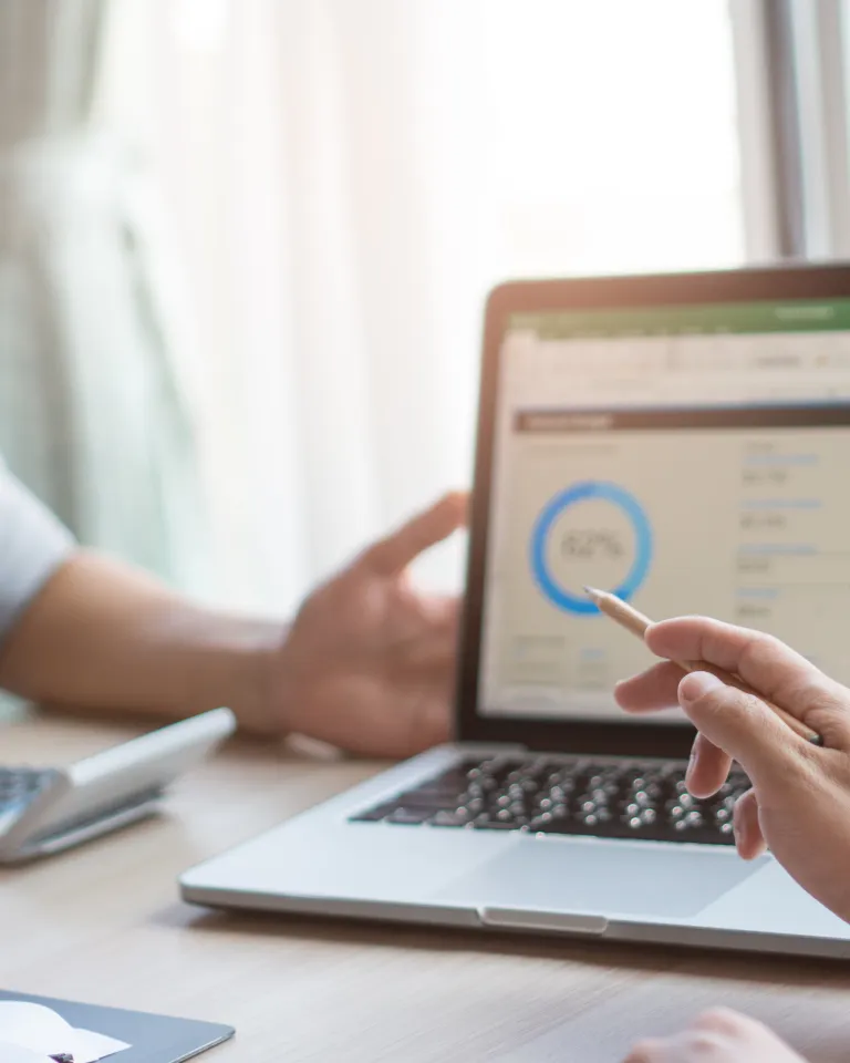 Two people discussing data displayed on a laptop screen, featuring a circular chart showing 62% and a bar graph. A hand points at the screen with a pencil. A calculator and papers are on the table.