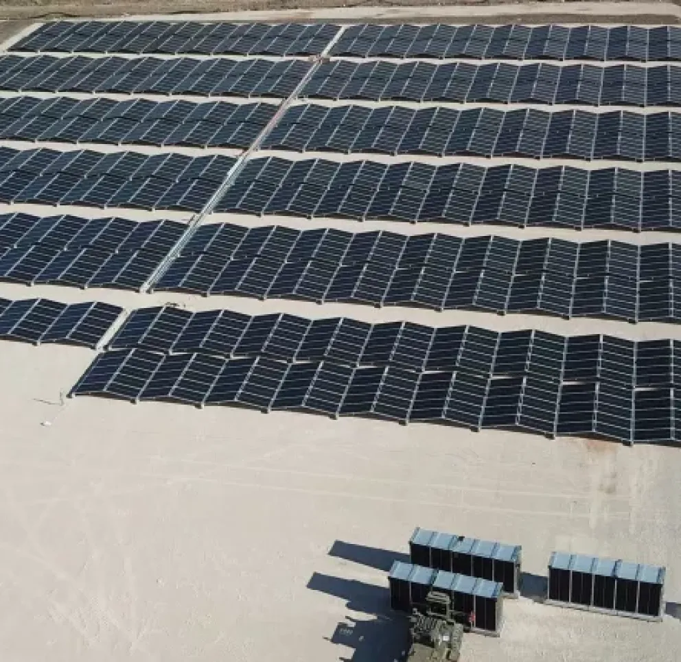 Aerial view of a large solar farm with multiple rows of solar panels on a flat, sandy terrain. The panels are arranged in a grid pattern, and there are small structures or equipment near the bottom of the image.