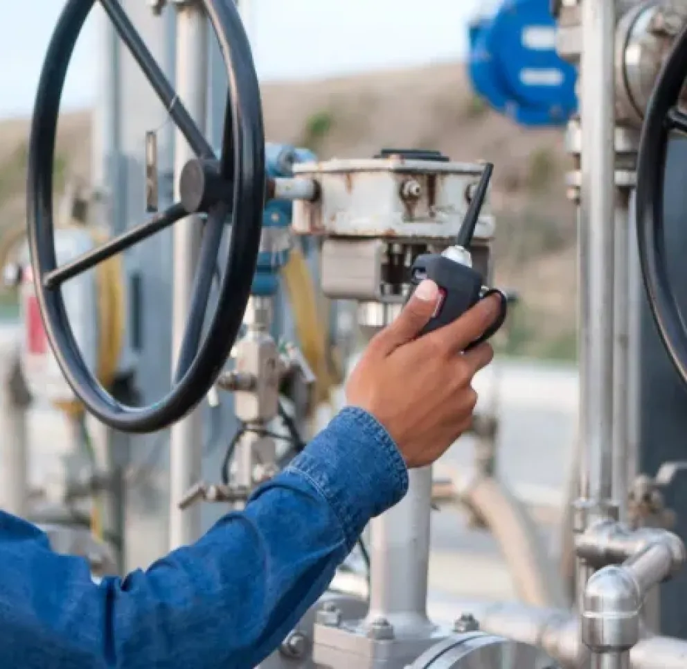 A worker wearing a hard hat and safety glasses operates a large industrial valve. The worker is holding a handheld device, possibly for communication or measurement. The setting appears to be an industrial site with metal pipes and equipment visible in the background.