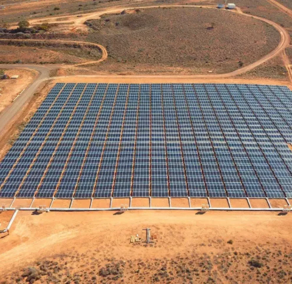 Aerial view of a large solar panel farm in a desert area. The panels are arranged in a grid pattern on a sandy terrain, surrounded by dirt roads. A small white building is visible at the bottom left corner.
