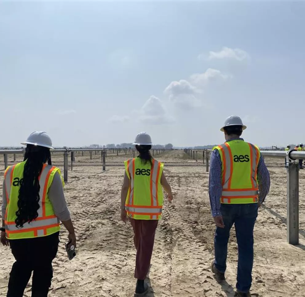 Three people wearing hard hats and AES-branded safety vests walk through a large, open construction site under a clear sky. They are surrounded by metal structures and equipment, with more workers visible in the distance.