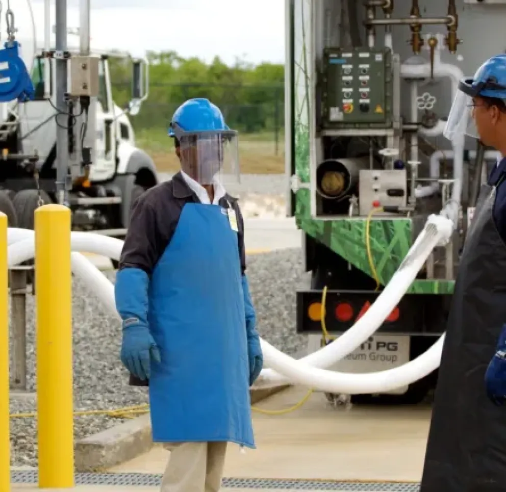 Two workers wearing blue helmets, face shields, gloves, and protective aprons stand near a tanker truck. They are surrounded by hoses and equipment, indicating an industrial setting. The background shows another truck and a fenced area.