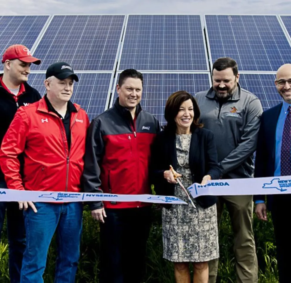 A group of people stand in front of solar panels during a ribbon-cutting ceremony. They are holding a ribbon marked 'New York State NYSERDA'. Two blue banners flank the group. The setting is outdoors with a clear sky.