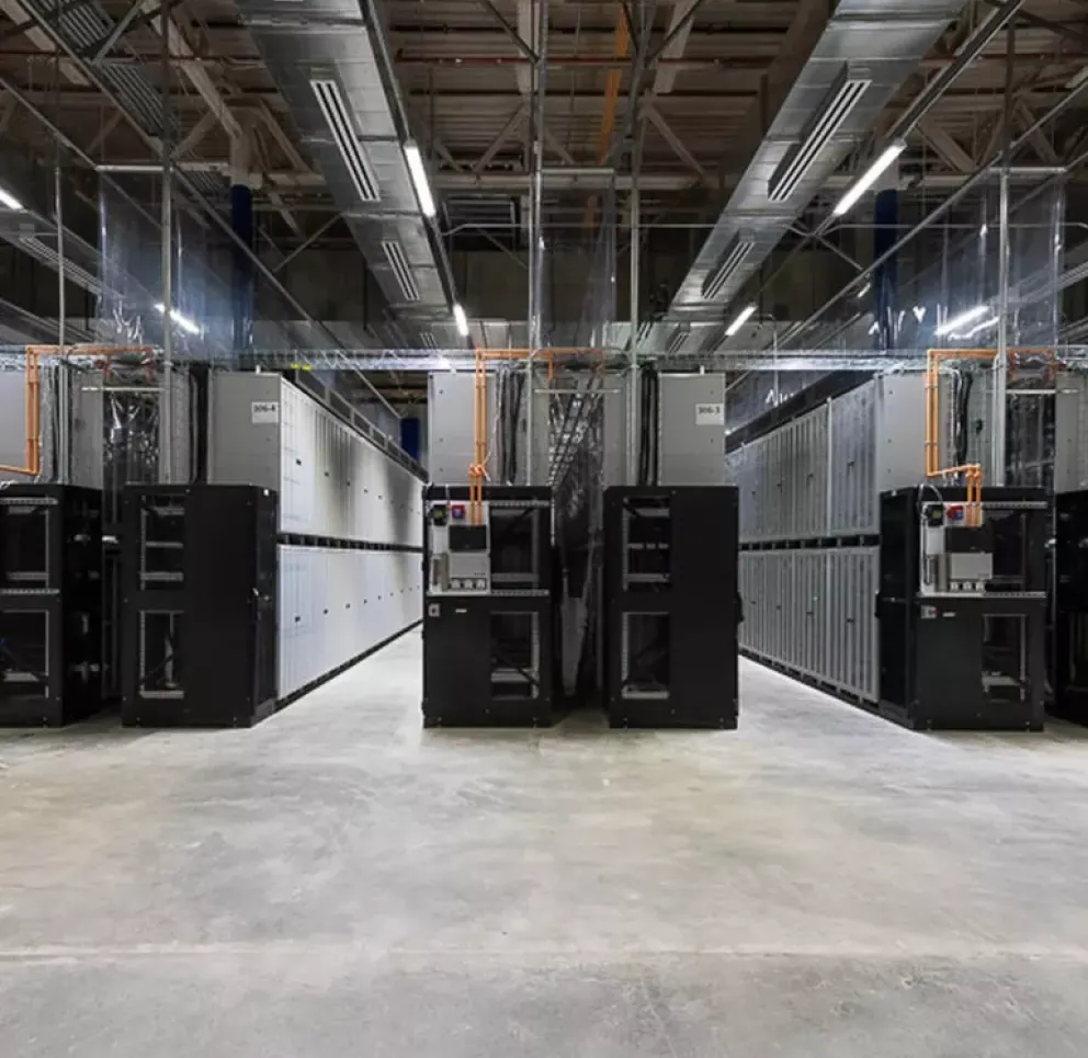 Interior of a large data center with rows of server racks under a high ceiling. The floor is concrete, and cables are visible overhead.