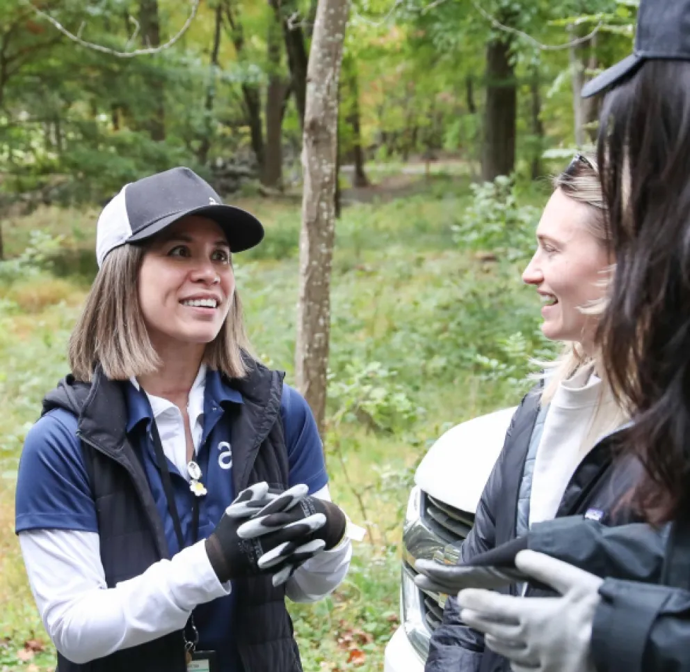 Three people in casual outdoor clothing, including gloves and hats, are having a conversation in a forested area. They are smiling and appear to be engaged in a friendly discussion.