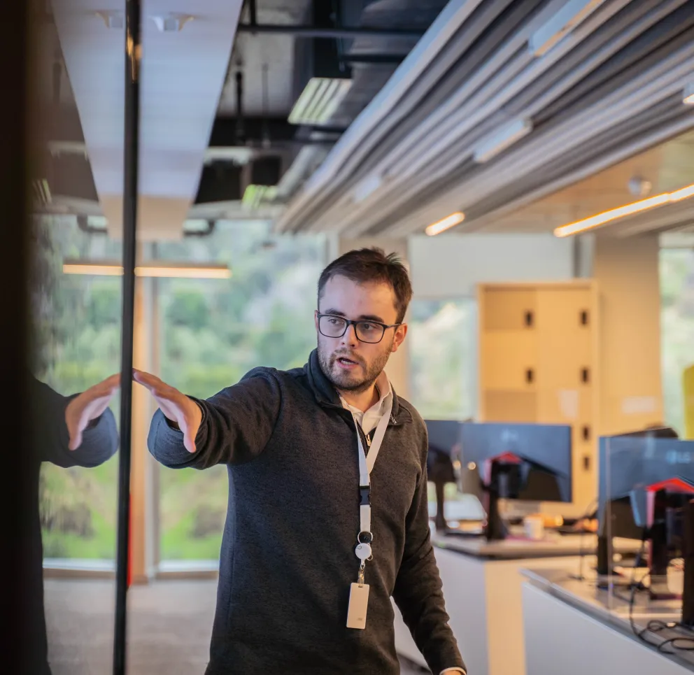 A man wearing glasses and a lanyard points to a reflective surface in a modern office with multiple computer monitors.