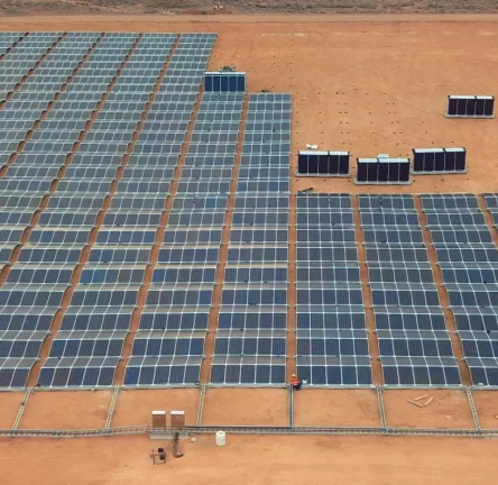 Aerial view of a large solar panel farm on a barren, sandy landscape. Rows of solar panels are aligned in a grid pattern, with several dark rectangular structures at the back. A small road is visible in the top left corner.