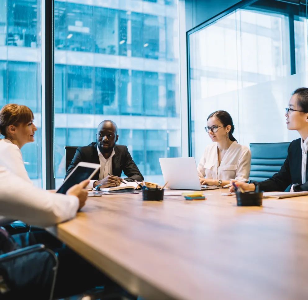 A diverse group of five professionals having a meeting in a modern conference room with large windows. They are seated around a wooden table, using laptops and tablets.
