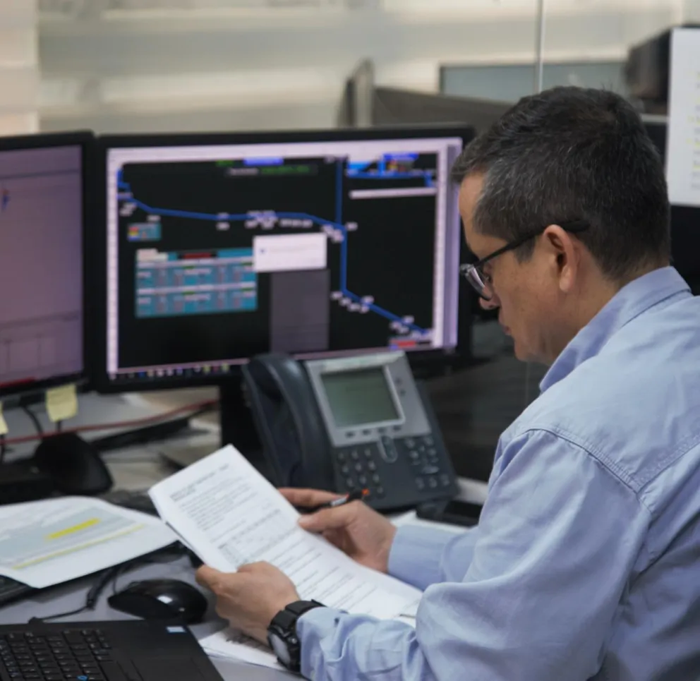 A person in a blue shirt reviews documents at a desk with multiple computer monitors displaying graphs and data. A phone and a laptop are also on the desk.