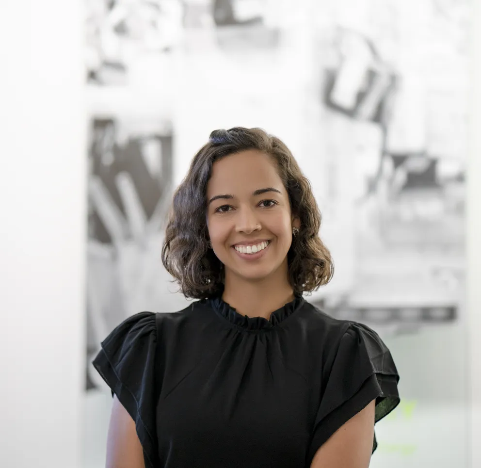 A person with shoulder-length curly hair wearing a black blouse smiles at the camera. The background is blurred with abstract black and white shapes.