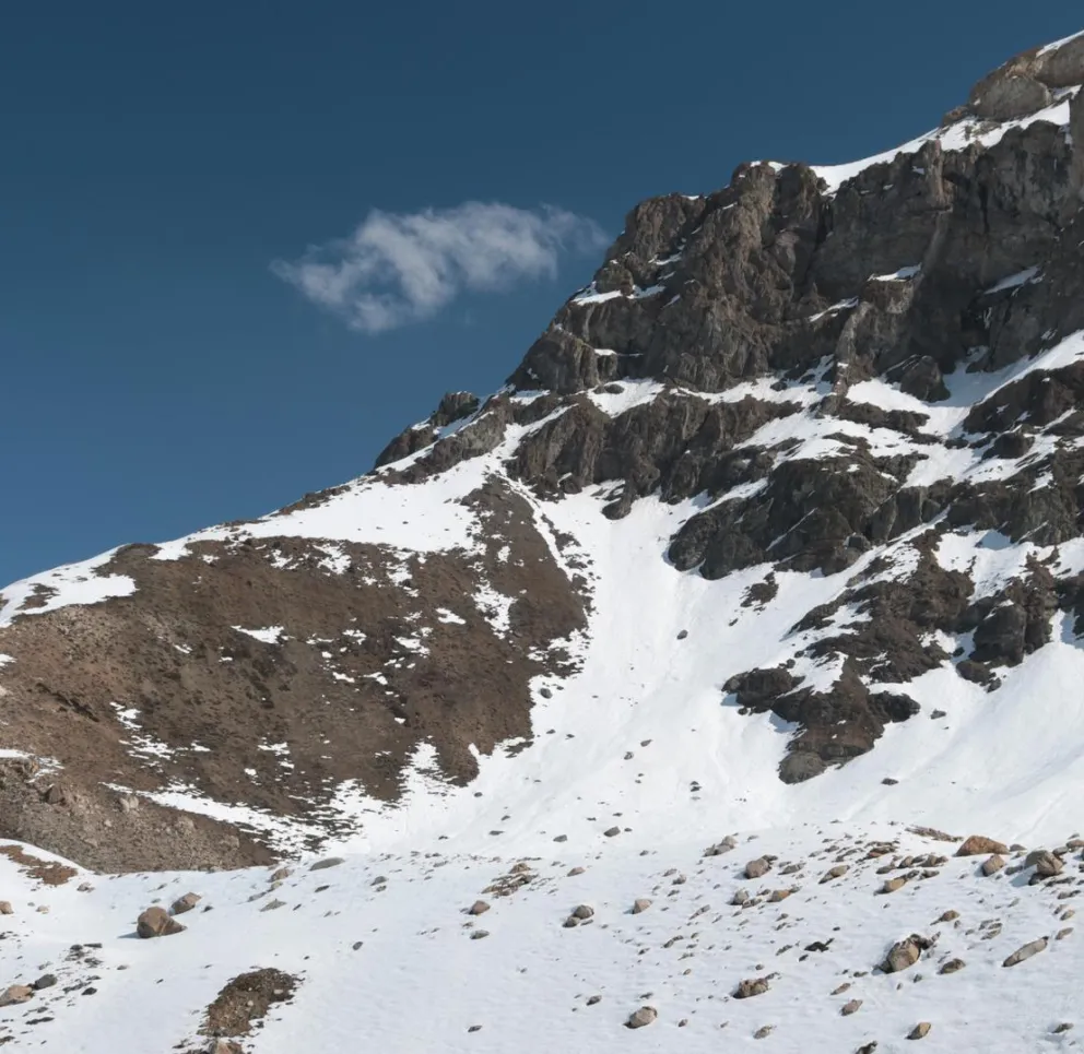 Snow-covered mountain slope with rocky outcrops under a clear blue sky. Sparse patches of snow reveal brown earth and rocks.