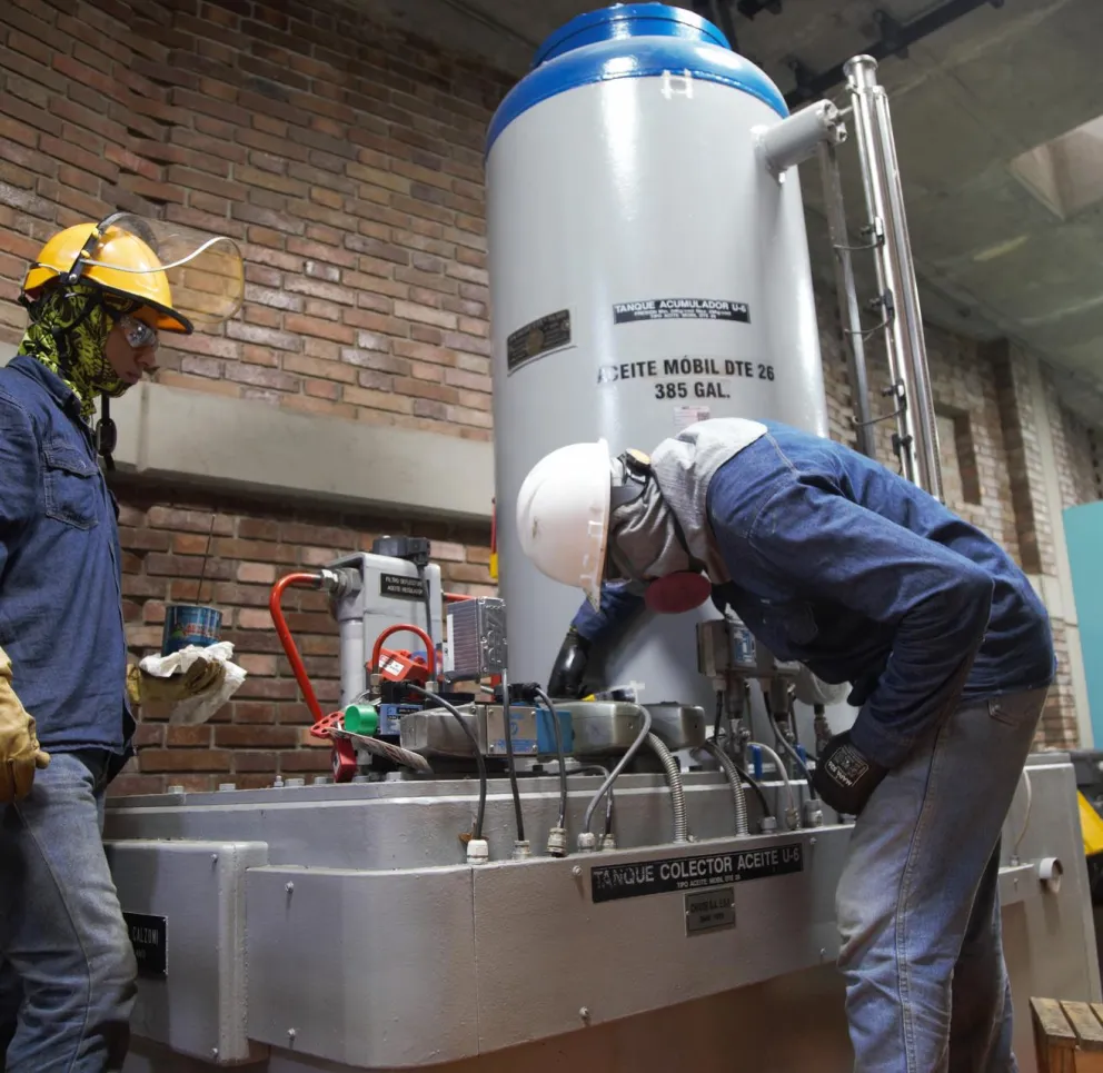 Two workers in protective gear operate machinery in an industrial setting. One is wearing a yellow hard hat and gloves, while the other leans over a large metal tank with a white helmet and respirator. The background features a brick wall and control panels.