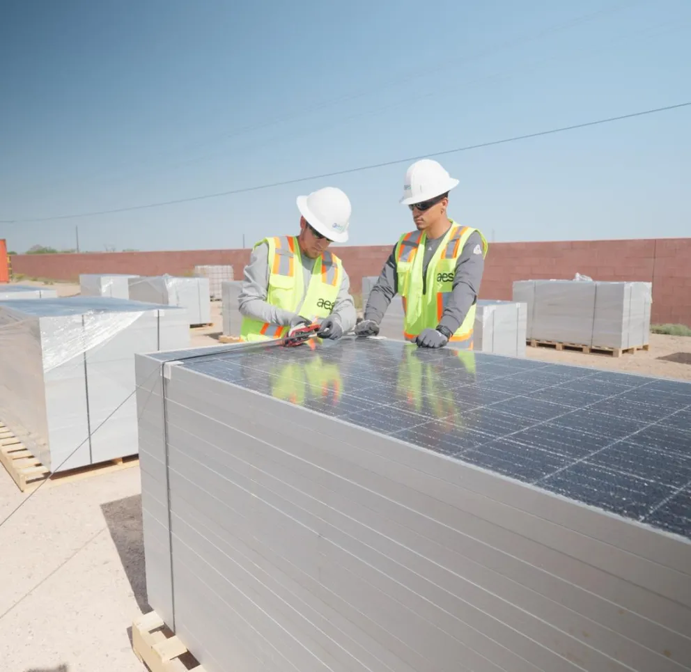 Two workers wearing safety gear inspect solar panels stacked on pallets in an outdoor area with a blue sky.