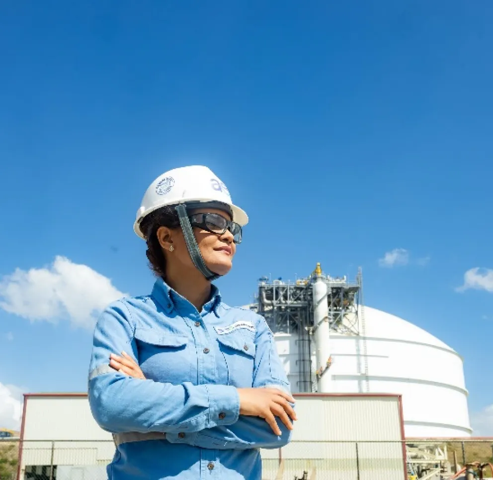 A person wearing a hard hat and safety glasses stands with arms crossed in front of a large industrial tank under a clear blue sky.
