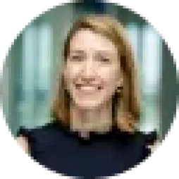 A person with shoulder-length hair smiling, wearing a dark top, in a blurred indoor setting.