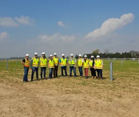 A group of people wearing hard hats and reflective vests stand in an open field with scattered metal posts. The sky is clear with a few clouds. Trees and a fence are visible in the background.
