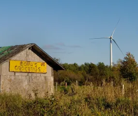 An old wooden building with a yellow sign that reads 'Bova's Groceries' stands in a grassy field. A large wind turbine is visible in the background against a clear blue sky.