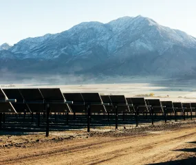 Rows of solar panels on a dirt field with snow-capped mountains in the background under a clear sky.